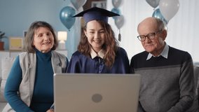 family with female graduate in hat and gown are listening to congratulations online via video communication on laptop while sitting at home in living room background of balloons