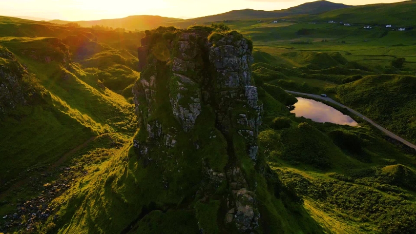 Landscape on the Isle of Skye in Scotland in the evening at sunset. Grassy hilltop Castle Ewen from Fairy Glen in deep sun. Clouds in the sky. Road with moving car meanders through the landscape. 