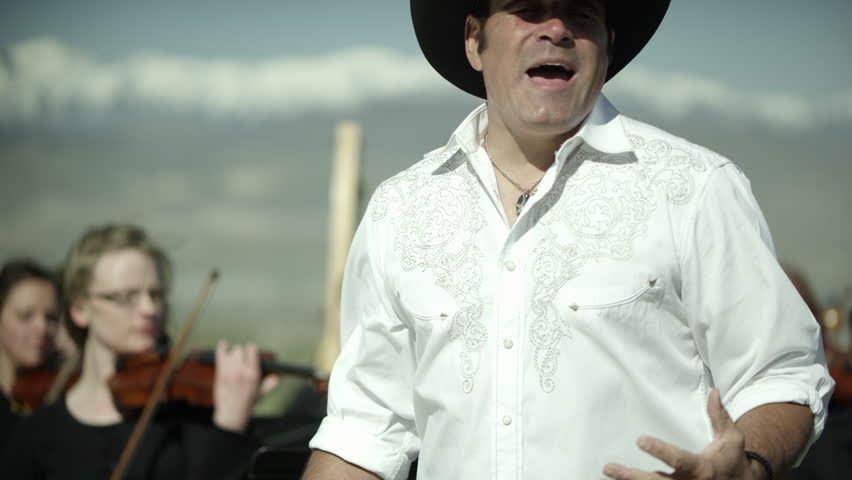 Country looking singer singing with outdoor orchestra in the mountains.