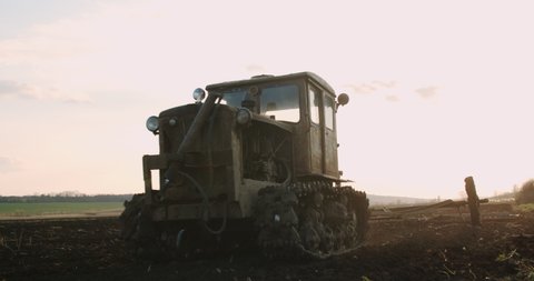 An old and rusty tractor, plowing the agricultural land, sunset. Slow motion.
