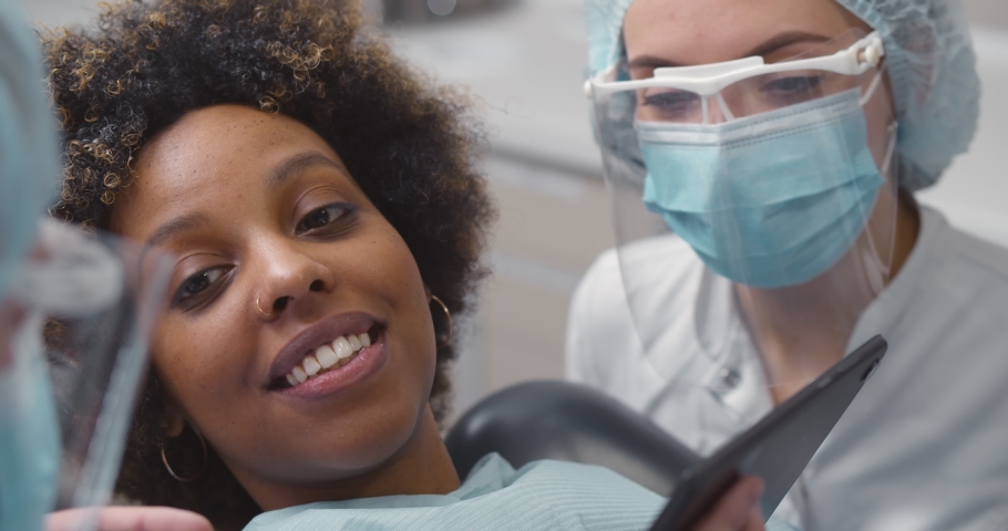Close up of dentist and assistant showing tablet computer to woman patient at dental clinic office. Orthodontist and nurse in protective mask showing teeth x-ray to african female patient | Shutterstock HD Video #1071731608