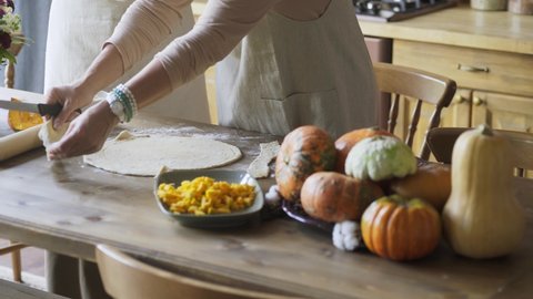 Unrecognizable old woman in white apron close-up hands circle and cut handmade dough with knife preparing festive thanksgiving pumpkin pie on kitchen in country style