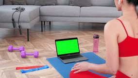 Green screen mock up chroma key monitor laptop concept: Fit woman in sportswear sits on yoga mat looks computer display, tells listen fitness trainer online video call webcam, distance sports lesson