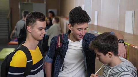Closeup of two tall teenage hooligans mocking smaller classmate in school locker room, pinching and pushing him aside. Grinning teenagers bullying younger schoolboy in school corridor