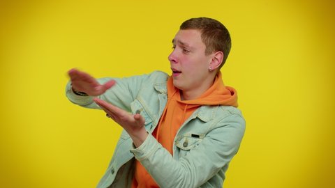 Cheerful rich teen stylish boy showing wasting or throwing money around hand gesture, more tips dreaming about big profit body language. Young man posing isolated on yellow studio wall background