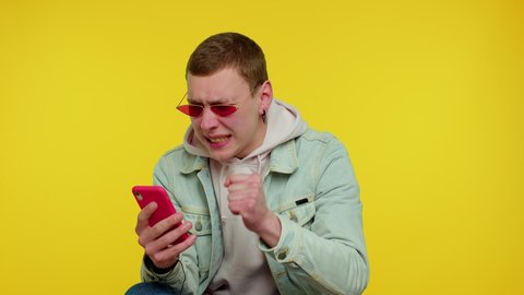 Teen stylish boy in denim jacket use mobile cell phone typing browsing, loses becoming surprised sudden lottery results, bad fortune, loss. Young man on yellow background. People sincere emotions