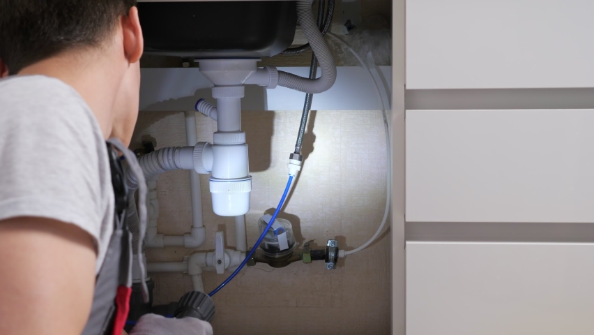 Plumber with glowing bright flashlight searches leakage on pipes and waste trap under sink in contemporary kitchen close backside view | Shutterstock HD Video #1071747448