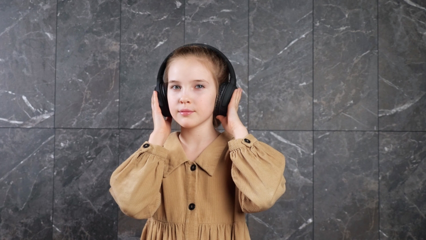 Young lady in brown clothes and black headphones listens to music and dances standing against decorative wall at school during break