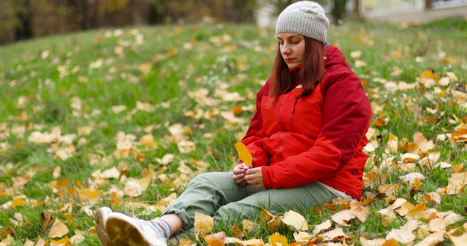 Young beautiful caucasian girl in warm clothes and hat sits on the grass with fallen leaves in the park. Girl hand holding a fallen leaf