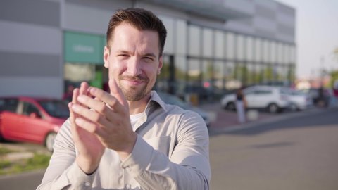 A young Caucasian man applauds to the camera and nods with a smile in an urban area - a supermarket in the blurry background