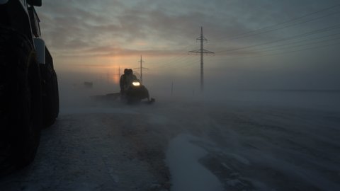 Snowmobile rides on icy road. Strong wind. Snowstorm. The setting sun. Snowfall. Power poles along the road. Snowfall