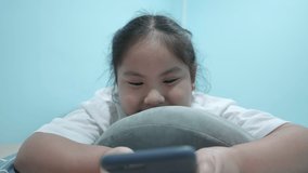 Happy chubby cute Asian girl watching online video, video call, message, cartoon, play game in smartphone. Adorable kid lie prone on pillow in home was smiling and laugh a lot. Front view