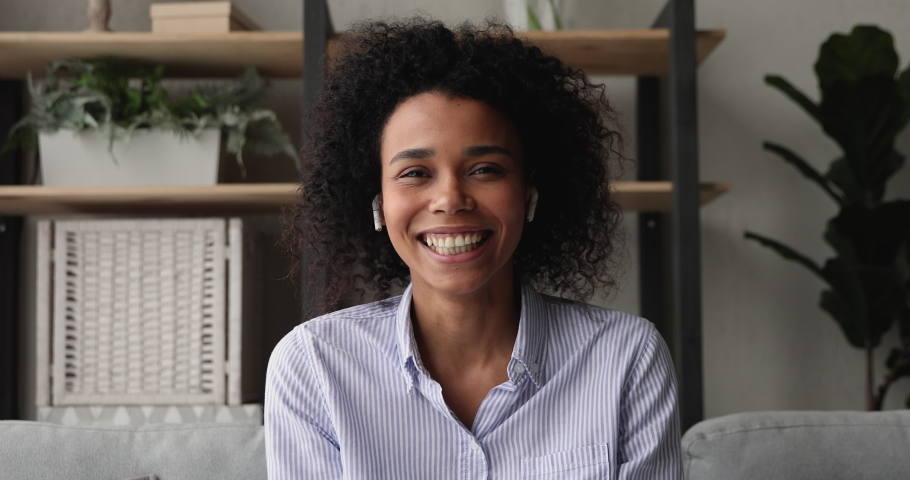 Head shot portrait African woman wear wireless earphones talk on videoconference, sit indoor look at camera greet friend having pleasant remote personal formal conversation. Video call event concept Royalty-Free Stock Footage #1071788455
