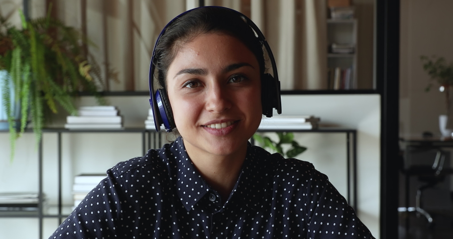 Head shot smiling pretty millennial indian ethnicity woman in headphones looking at camera, involved in distant video call zoom conversation, holding project negotiation remote meeting with colleagues Royalty-Free Stock Footage #1071788476