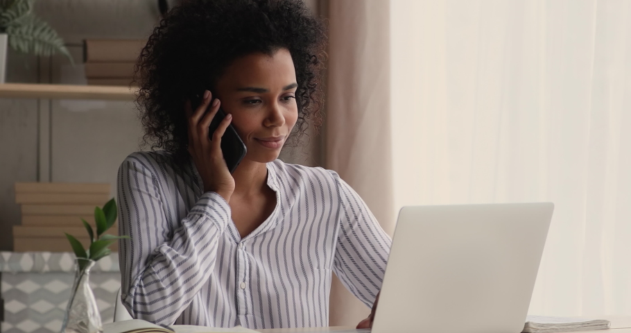 African businesslady talk to client on cellphone use laptop discuss deal, millennial woman sit at desk at home look at computer screen makes order remotely receive consultation support by phonecall Royalty-Free Stock Footage #1071788479