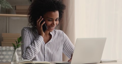 African businesslady talk to client on cellphone use laptop discuss deal, millennial woman sit at desk at home look at computer screen makes order remotely receive consultation support by phonecall