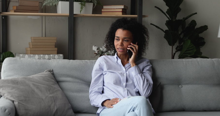 Smiling young African ethnicity woman talk on the phone. Attractive female spend day off at home sit on sofa in living room alone having pleasant remote conversation using modern tech and connection Royalty-Free Stock Footage #1071788485