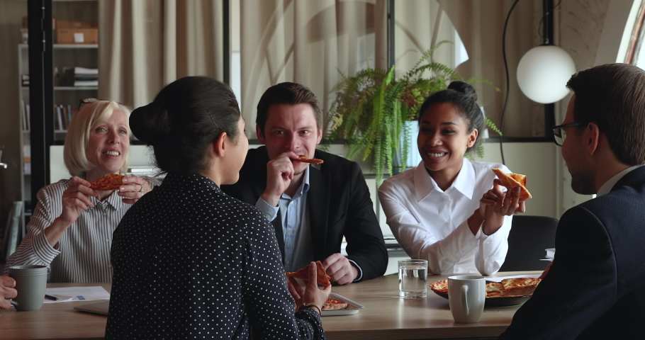 Happy multiracial older young diverse business people colleagues sitting at table, enjoying communicating chatting relaxing during break pause time eating tasty italian pizza in modern office room. Royalty-Free Stock Footage #1071788512