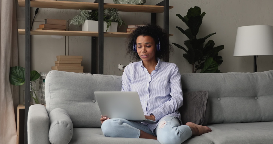 African woman resting on couch at home wear wireless headphones using laptop talk to family or friend on videoconferencing application, pleasant personal remote conversation. Video call event concept Royalty-Free Stock Footage #1071788593