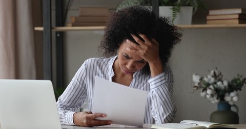 African woman sit at workplace desk holding papers reading bad news in letter feels frustrated concerned due high taxes, loan debt, dismissal, staff cuts notice, debt eviction notification concept