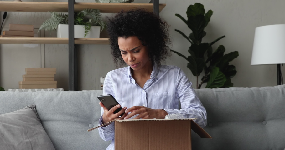 Young African woman sit on sofa opens parcel box check ordered goods in shopping app on cellphone getting wrong damaged items feels dissatisfied leaves negative feedback. Bad delivery services concept Royalty-Free Stock Footage #1071788701