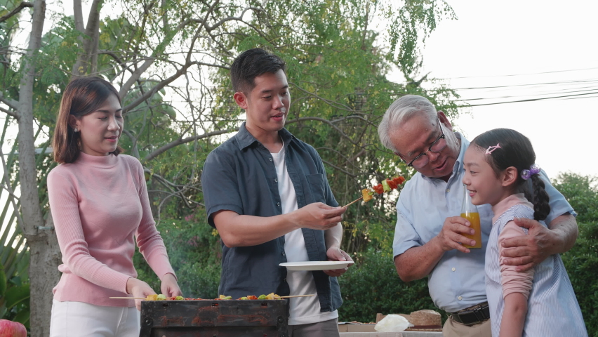 Happy Asian family having a barbecue-BBQ party together in the garden at home. New normal family lifestyle and happy family funny activity at home