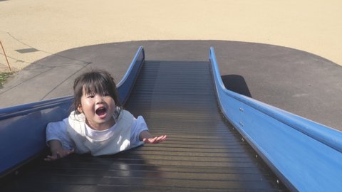Funny Asian little toddler girl playing on the slide in prone position. Playground in the park. Happy smiling 3 years old child. Motion of Gimbal shot. Lifestyle, parenting, leisure, outdoor, activity