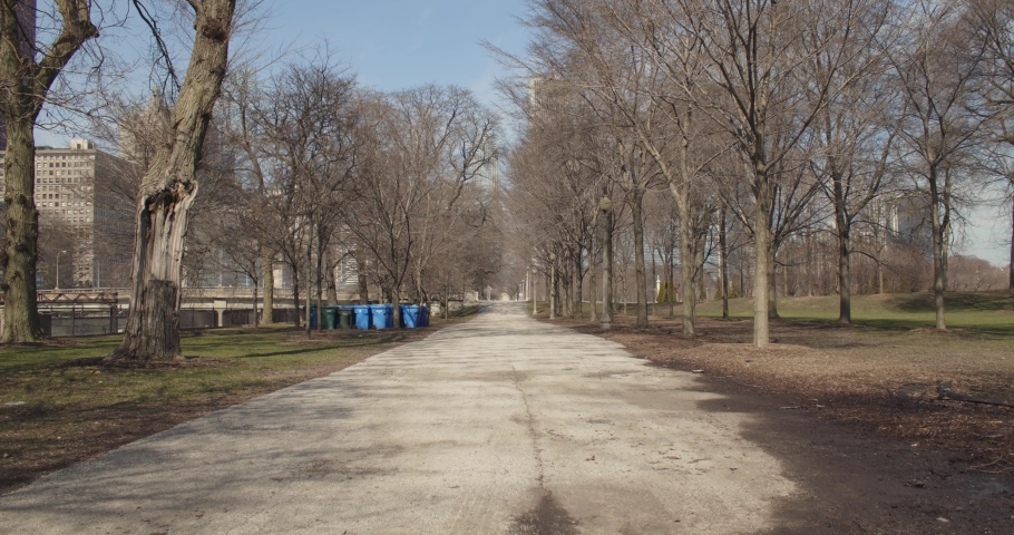 Fall lock-down footage of Grant park in Chicago, Illinois. Looking north over dormant trees and a dirty asphalt walkway. Trash and recycling cans in the distance with urban life in the background.