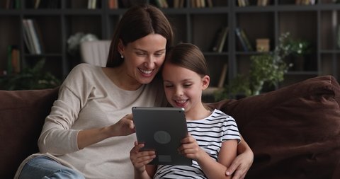 Loving mom and little daughter sit together on sofa at home with tablet device choose buy goods on internet, using education application for easy and funny learning, modern tech and generation concept