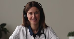 Head shot 35s female GP doctor wear white coat and headset looks at camera communicates to patient remotely. Provide professional help, support, give advices. On-line consultation, video call concept