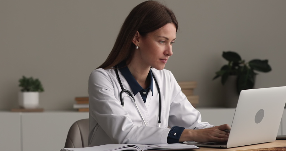 Young woman general practitioner in white coat stethoscope on neck working on laptop writes notes filling medical journal while sit at workplace desk. Busy medical worker, workday in clinic concept Royalty-Free Stock Footage #1071802543