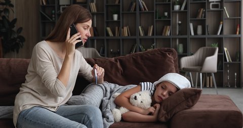 Concerned young mother using digital thermometer measures temperature of sick daughter talk by phone receive medical help remotely, unhealthy kid girl with towel on head hug fluffy toy lying on sofa
