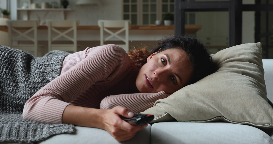 Idler young woman lying on couch covered with plaid at home alone holding TV remote control switch channels waste time feels bored. Lazy day off at home, unmotivated female, inactive lifestyle concept Royalty-Free Stock Footage #1071802639