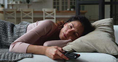 Idler young woman lying on couch covered with plaid at home alone holding TV remote control switch channels waste time feels bored. Lazy day off at home, unmotivated female, inactive lifestyle concept