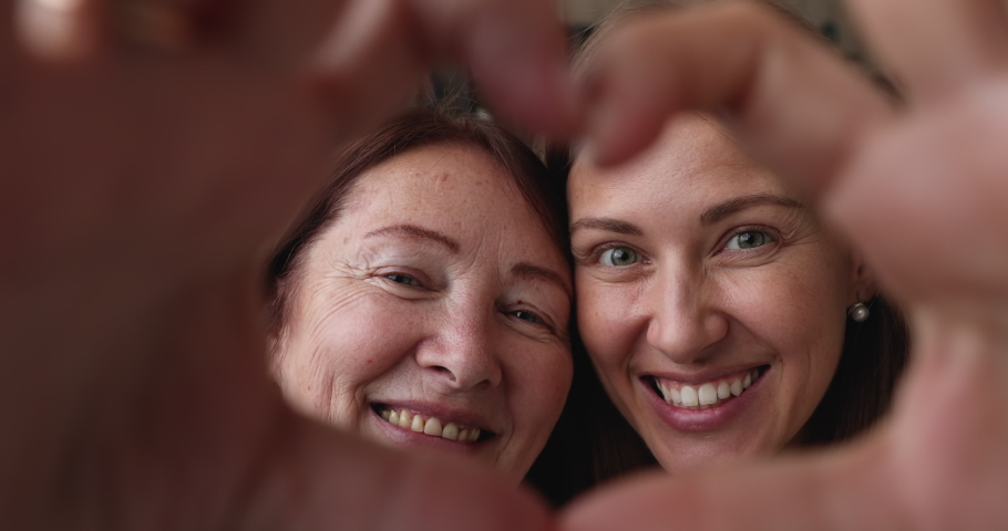 Close up older mom and grownup daughter happy faces seen through joined fingers making heart gesture, multigenerational family smiling look at camera feeling love enjoy moment. Bond, affection concept