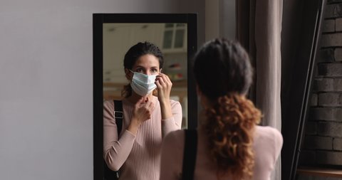 Young woman before leaving home put on surgical face mask standing in front of mirror in hallway, facemask mandatory in all public places due pandemic outbreak of covid. Personal safety, care concept