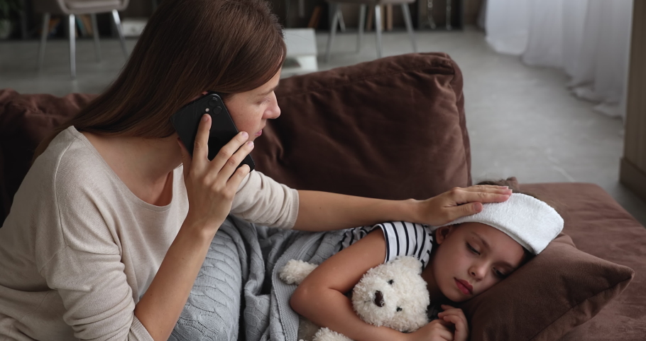 Concerned mother talk on phone with therapist stroke touch her sick little daughter while she lying on sofa with towel on head, mom receive medical aid distantly, kid has coronavirus symptoms concept Royalty-Free Stock Footage #1071802834