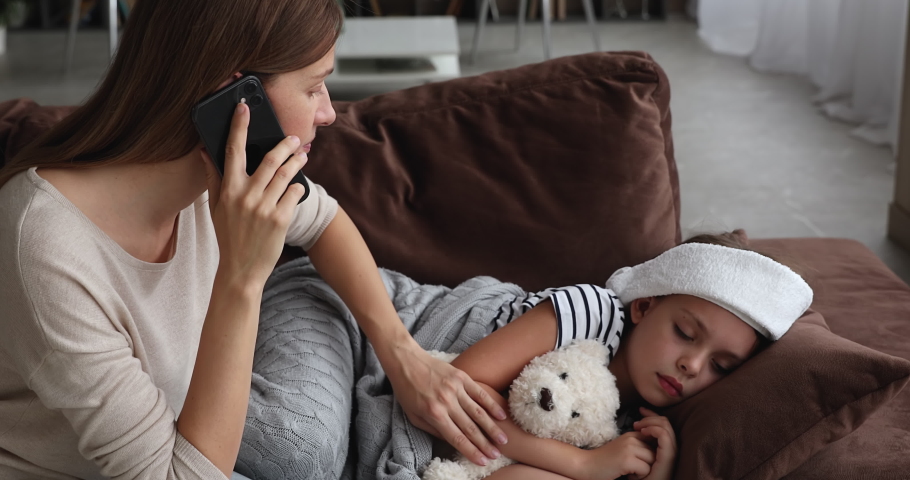 Concerned mother talk on phone with therapist stroke touch her sick little daughter while she lying on sofa with towel on head, mom receive medical aid distantly, kid has coronavirus symptoms concept | Shutterstock HD Video #1071802834