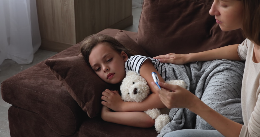 Young woman holding digital thermometer put hand on kid head check temperature caring of little lovely daughter lying on sofa with fluffy toy, corona virus symptoms, need medicare, medications concept | Shutterstock HD Video #1071802861