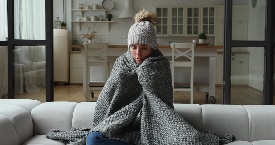 Young woman wear hat wrapped in knitted plaid sit alone shivering from cold on sofa in unheated apartment without central heating due debt. Unhealthy female feeling discomfort try to warming up indoor Royalty-Free Stock Footage #1071802882