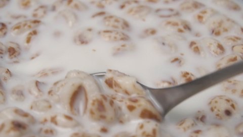 Spoon stirring instant dairy puffed spelt wheat breakfast in slow motion. Mixing and cooking cereal flakes with milk. Macro shot