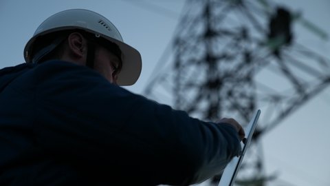Power engineer in protective white helmet checks power line online using computer tablet, remote access to energy system, for control and safety. High voltage electrical lines at sunset. clean energy