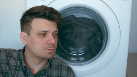 Tired man is waiting the washing machine with grey bedspread inside it, doing his laundry at home or laundry room. Everyday life, housework and household of young man.