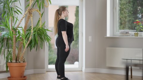 Side view of motivated overweight sportswoman doing jump squats indoors. Wide shot of confident Caucasian young woman training at home. Fitness and healthy lifestyle concept