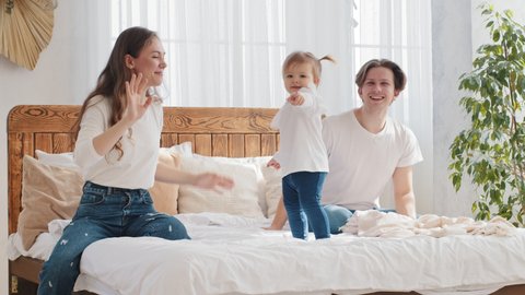Family having fun at home bedroom in morning, little girl toddler dancing to music on bed father and mother support rejoice enjoying daughter dance child points with finger to camera mom waving hello