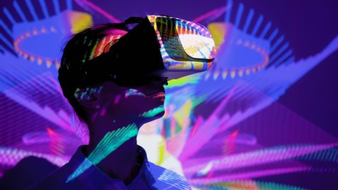 Woman using virtual reality headset and looking around at interactive technology exhibition with changing colorful projector light illumination. VR, augmented reality, immersive, entertainment concept