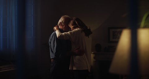Cinematic shot from window of happy mature senior couple in love is enjoying time together and dancing to celebrate their timeless love at home in evening. Concept: love, family, marriage, home, age.