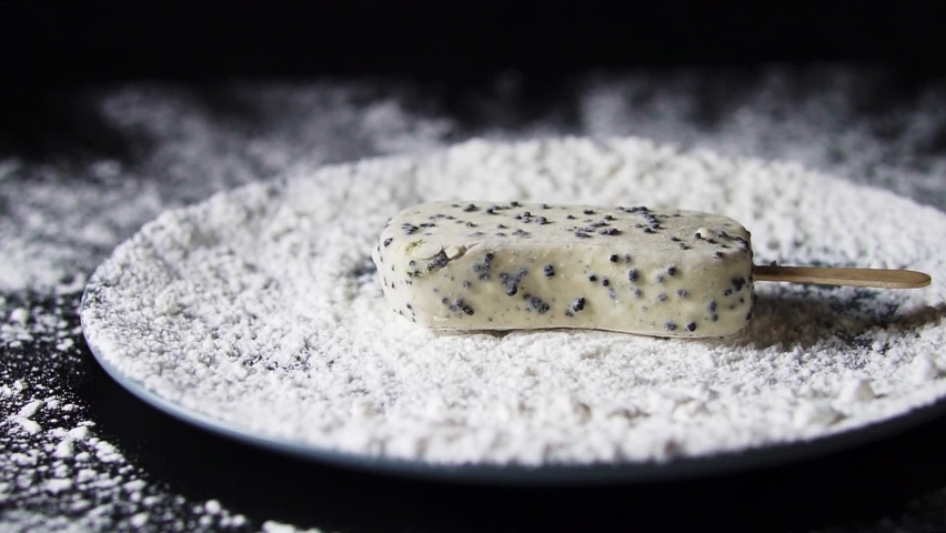White ice cream sundae falls into white powder in a plate. Powdered sugar is scattered over a black background. Falling chocolate into a plate
