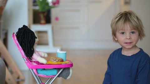 Little toddler child, boy, feeding his favorite doll, sitting in doll chair at home