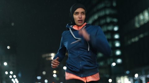 Cinematic shot of young arab sportswoman with athletic body in hijab running with effort and dedication in city center with snow falling at night. Concept:determination,woman power,goal achievement.